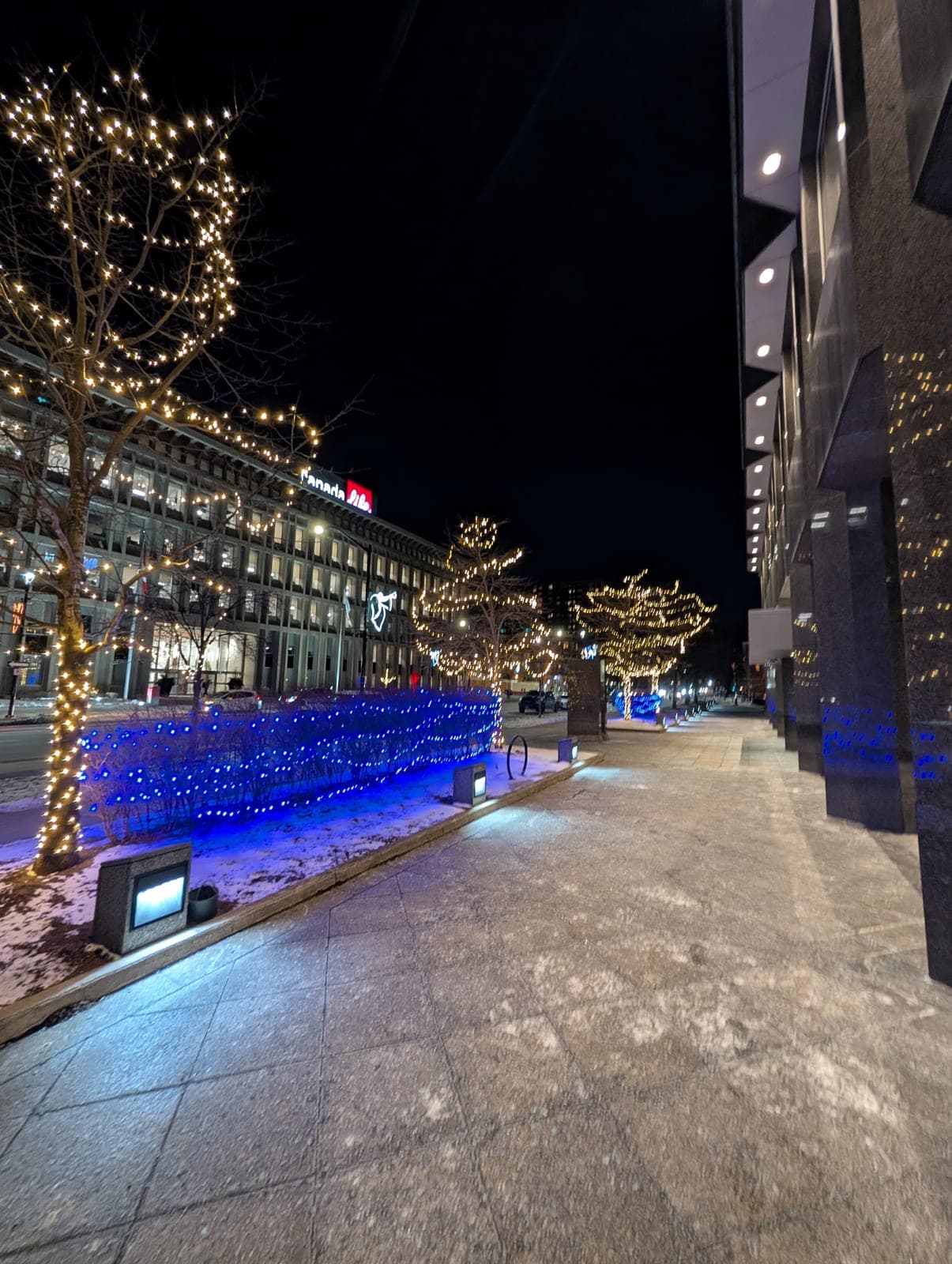 Snowy downtown sidewalk with warm-white tree wraps and blue landscape lighting beside a granite office tower.