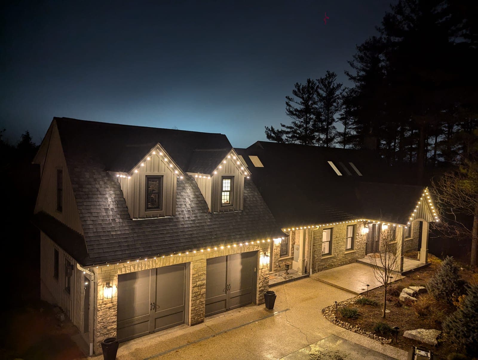 Stone and siding house with warm-white roofline Christmas lights against a deep blue evening sky
